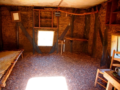 [Canvas on the ceiling keeps the sod from falling in the room. A cot is on the left and a table and chairs are on the right with a open square for a window in the middle.]